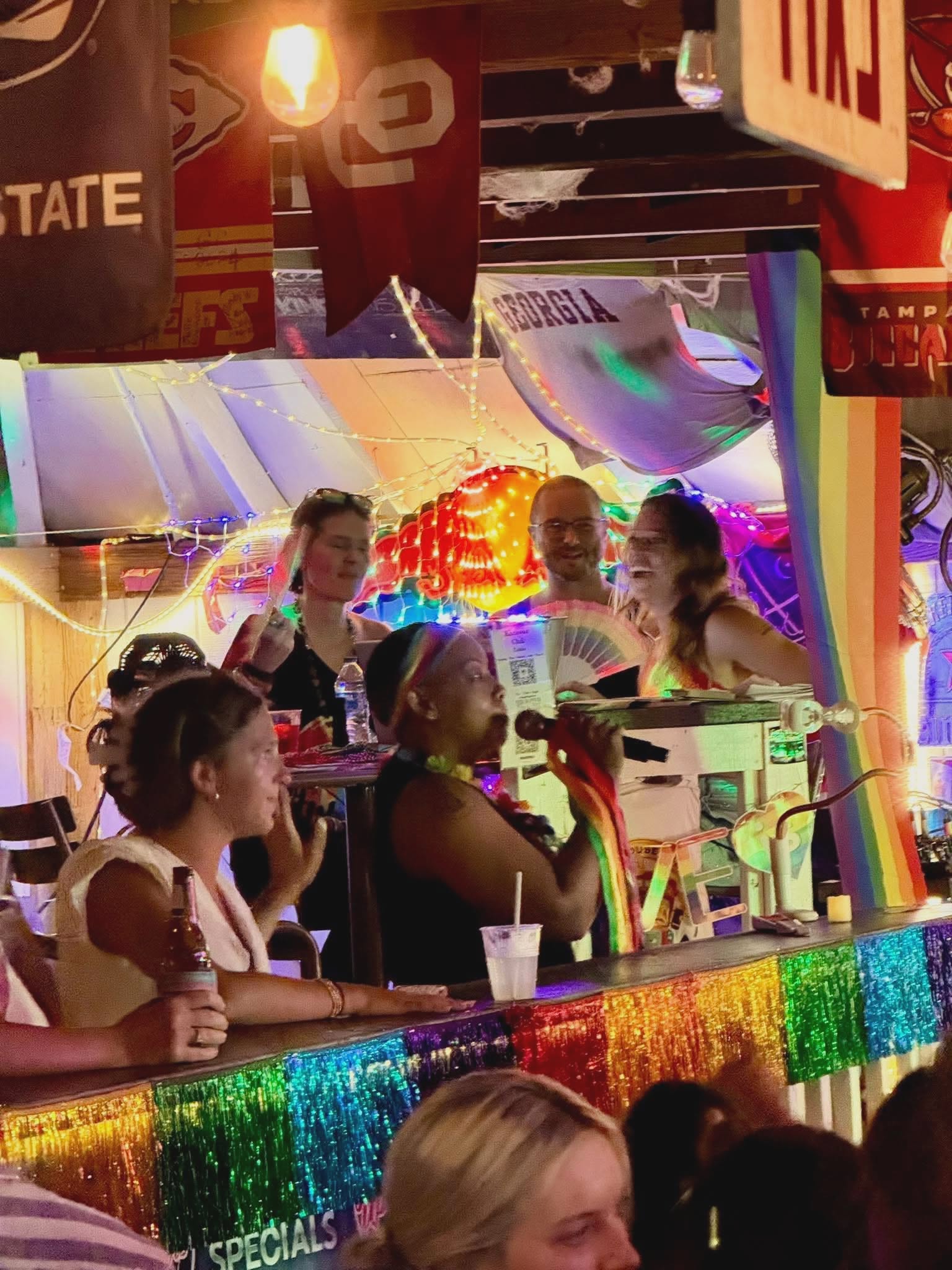 A singer on the mic while the crowd packs the bar under rainbow tinsel